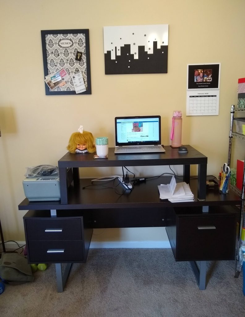 A standard office desk with three drawers. A matching coffee table is placed on top of it to make a standing desk. There is a computer on the desk, and we can see the author is clearly browsing Facebook rather than working. There appears to be facial tissues coming out of a creepy doll's head in the top left. WTF?