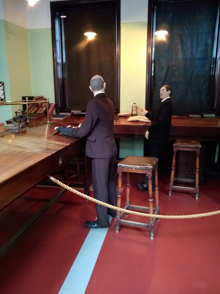 The preserved office of the Verdant Works factory museum. A standing-height ledge runs along two walls. Two mannequins dressed as old clerks are posed to look like they're going through the ledgers. Although there are two tall stools available, both men are standing without touching them. 