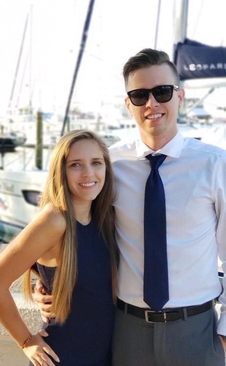 Amelia U, a chronic sciatica sufferer, is standing with her husband in a harbor. She is wearing a black dress and has her hand on her hip. He is wearing a white shirt, tie, and sunglasses. Both are smiling.