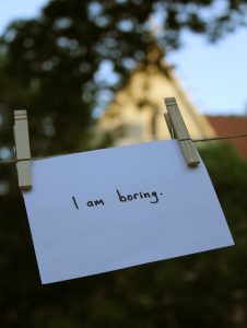 A note card is hung on a laundry line with clothespins. The sentence "I am boring" is written on it in marker.