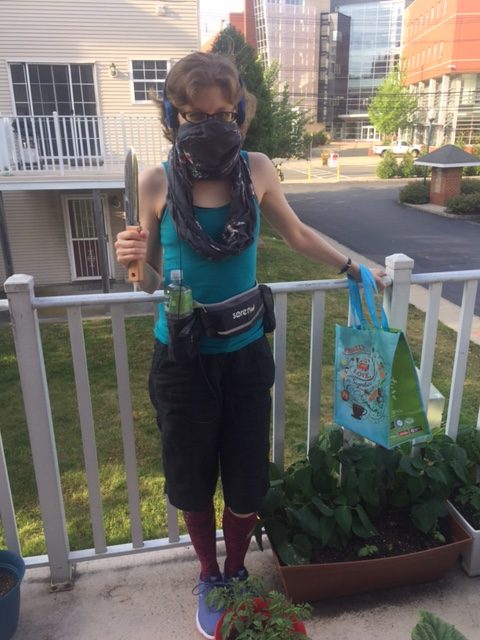 A photo of me in my typical going-out costume. I'm standing on my balcony, surrounded by plants. My face is half-covered by a scarf, in order to ward off the coronavirus. I am holding a foraging digging stick in one hand, and a brightly-colored reusable bag in the other. I am wearing a blue tank top, baggy black knee-length capris, knee-high compression socks, and blue tennis shoes.