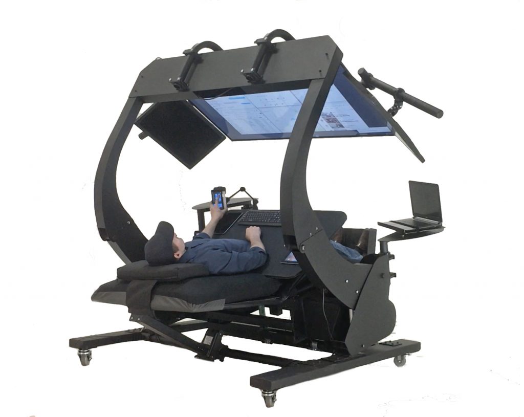 A photo of a man reclining in an Ergoquest workstation. The seat fully reclines, and arms wrap around from behind and dangle a large computer monitor in front of his face. He is touching his phone, which is supported on the left. A mount on the right holds a separate laptop.