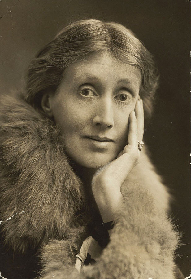 A sepia photo of Virginia Woolf from 1927. She is gazing straight into the camera, with a slight smile. Her left hand brushes her cheek. She is wearing a fur coat and a heavy ring.