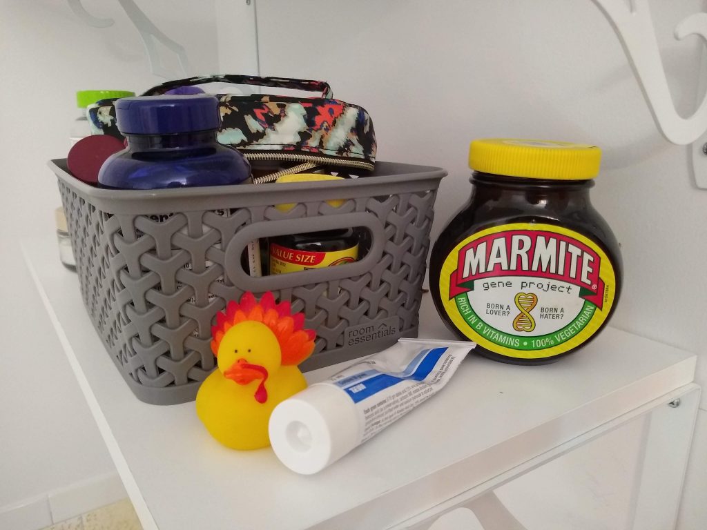 My medicine shelf. There is a gray plastic bin containing my pill bottles. In front is a tube of ointment, and a rubber ducky. To the right is a giant jar of Marmite.