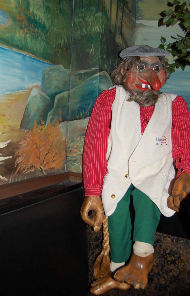 A carved wooden troll wearing a red shirt, white vest, and green pants stands in front of a mural of the Norwegian countryside.