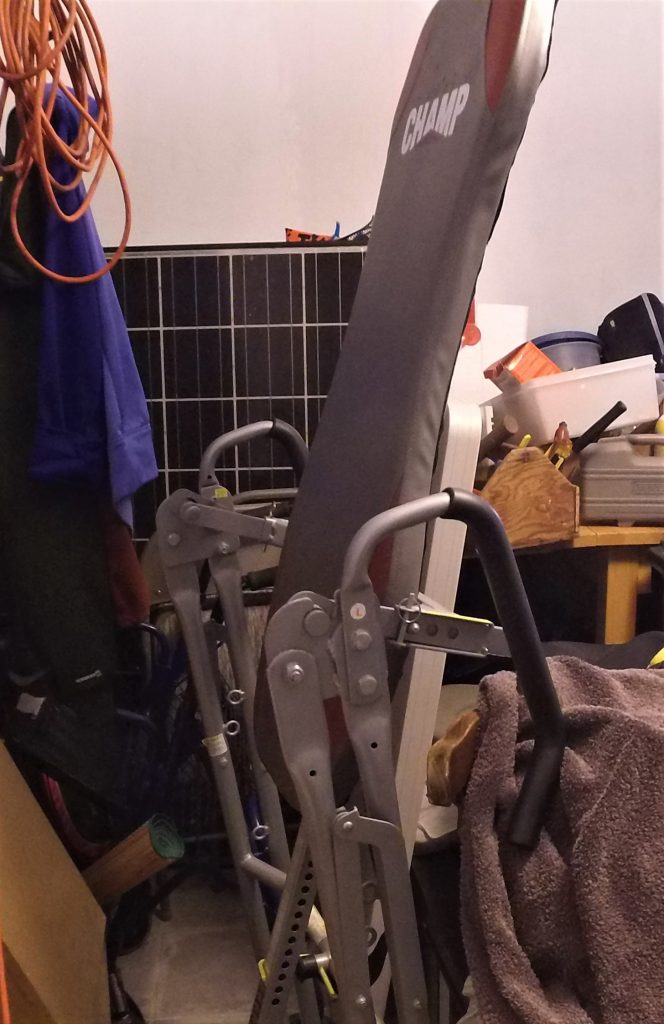 A photo of Stan's inversion table. It's folded up and crammed into a corner of a storage room.  A towel bumps into it on one side. Other boxes and loose objects are visible in the back.