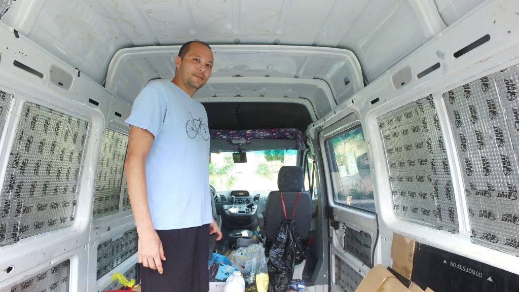 A photo of Stan standing inside his van. Stan is 6 feet tall, and able to stand upright. The floor is still littered with odds and ends, and the inside is clearly unfinished. A trash bag is strung over the back of the passenger seat.
