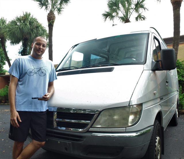 A picture of stand standing next to his white van. There are palm trees visible in the background.