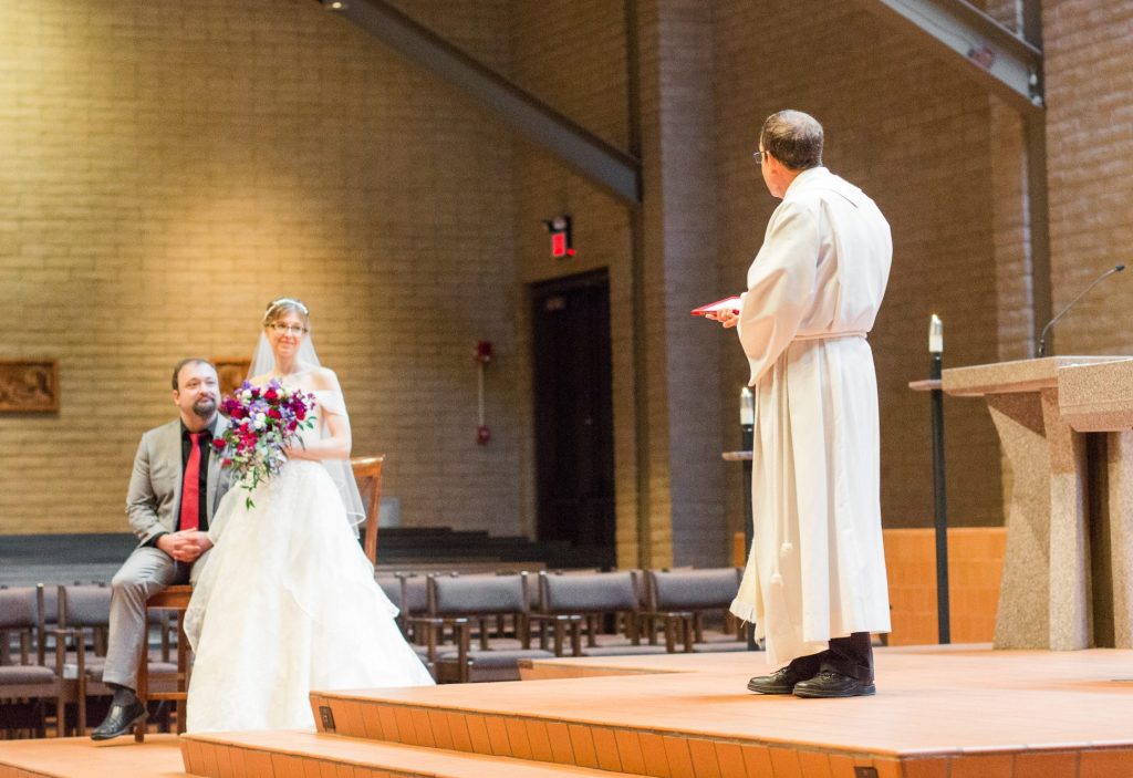 A photo from our wedding ceremony. My husband is sitting on a high chair on the left, while I stand on the right. The priest stands in the foreground, no doubt imparting some sacred wisdom upon us.