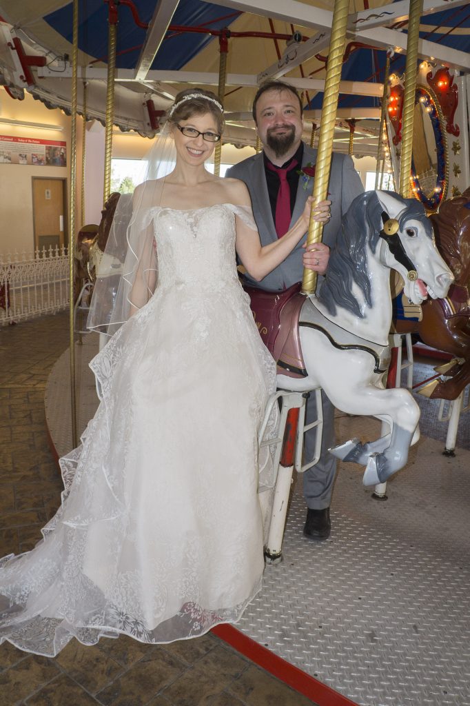 A photo of my husband and me taken at our wedding reception. Were standing by a white horse on a carousel. My husband is standing behind it, and Im standing in front in my poofy dress. Im awkwardly NOT SITTING DOWN!