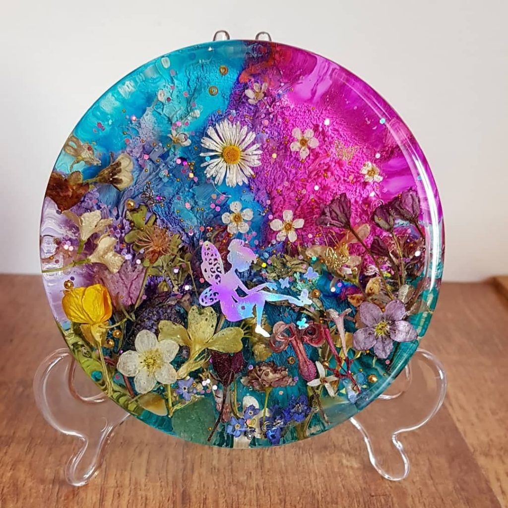 A "fairy meadow" crafted out of epoxy resin. Deb layers colored and clear resins with real flowers and seeds to create the effect. The silhouette of a seated fairy is visible in the middle. There are sparkles throughout the piece, which I am told come from fairy dust.
