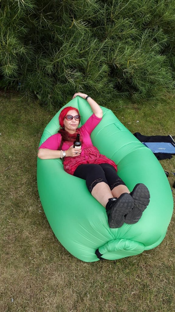 Deb kicks back on a large inflatable bean-bag type chair. She's outside. There are tree branches visible in the background, and the chair thing is set on the grass. Deb wears sunglasses and hiking shoes, and clutches a beer in her right hand.