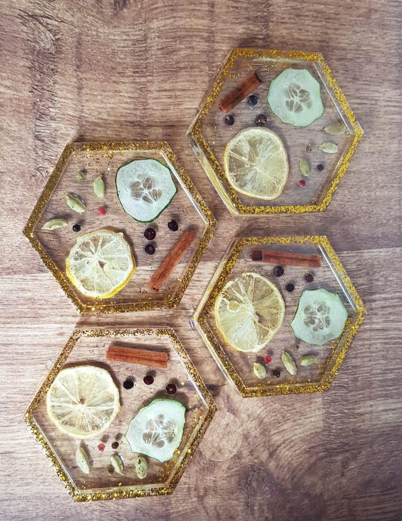 A set of four hexagonal coasters set on a wooden surface. The edge of each coaster is dusted in gold. Set inside the clear resin of each is a slice of lemon, a slice of cucumber, a cinnamon stick, and little seeds which may be black pepper and lemon pips.
