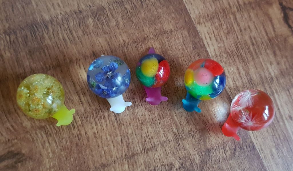 A group of five snails that Deb made out of epoxy resin. They are all set in a semi-circle on a wooden surface. Each snail is unique, three contain flowers or seeds, and two have mottled colored resins.