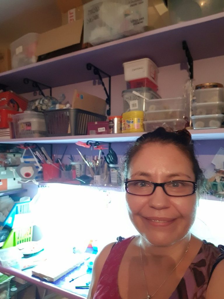 Deb stands in her workshop. Shelves full of supplies are visible above her head. Her work surface is visible behind her, but it's obscured due to the glare from a lamp.