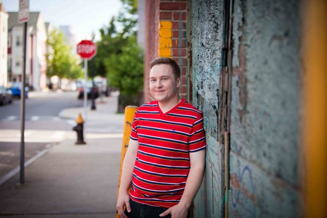 A photo of Nick taken in 2014. Nick is standing on a city street, leaning against a building. He's smiling and waiting a red shirt.