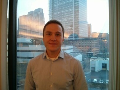 A photo of Nick taken in 2014. The photo shows Nick standing in front of the large window at his office. Skyscrapers are visible in the background. Nick is wearing a button-down shirt and is grinning.