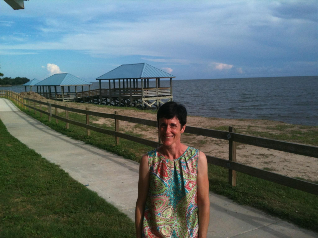 A photo of Claire besides the seashore in Louisiana. She's standing on the grass in front of a sidewalk. Behind the sidewalk is a wooden fence, and beyond that is the beach.  There's a wooden structure projecting into the ocean that looks like a water gazebo. Is it a water gazebo? What else would you call it? I'm from North Dakota, I don't know about water.