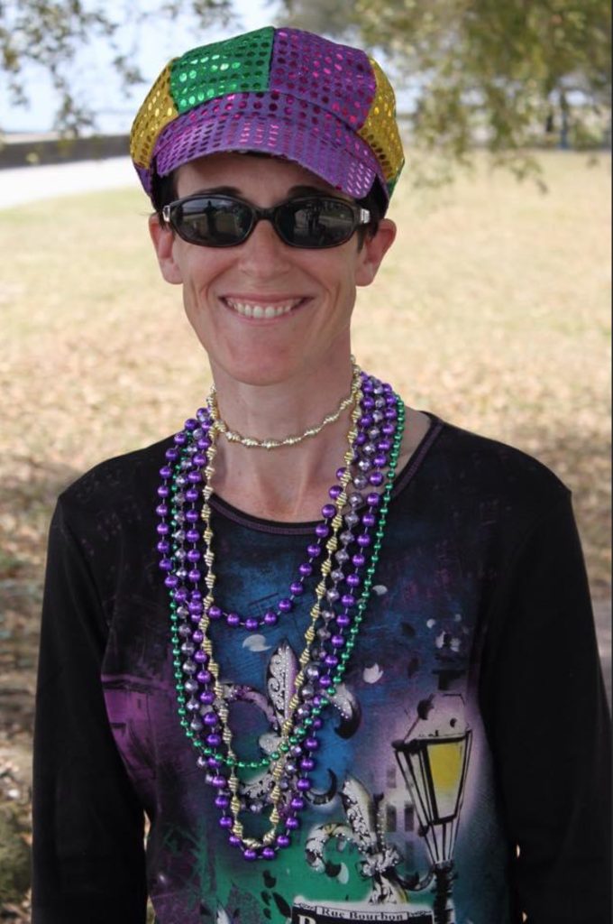 A portrait of Claire taken during Mardi Gras in 2014. She's wearing a garish hat, with yellow, green, and purple sequins. She's also wearing a half-dozen strands of Mardi Gras beads around her neck. She's wearing sunglasses, and smiling.