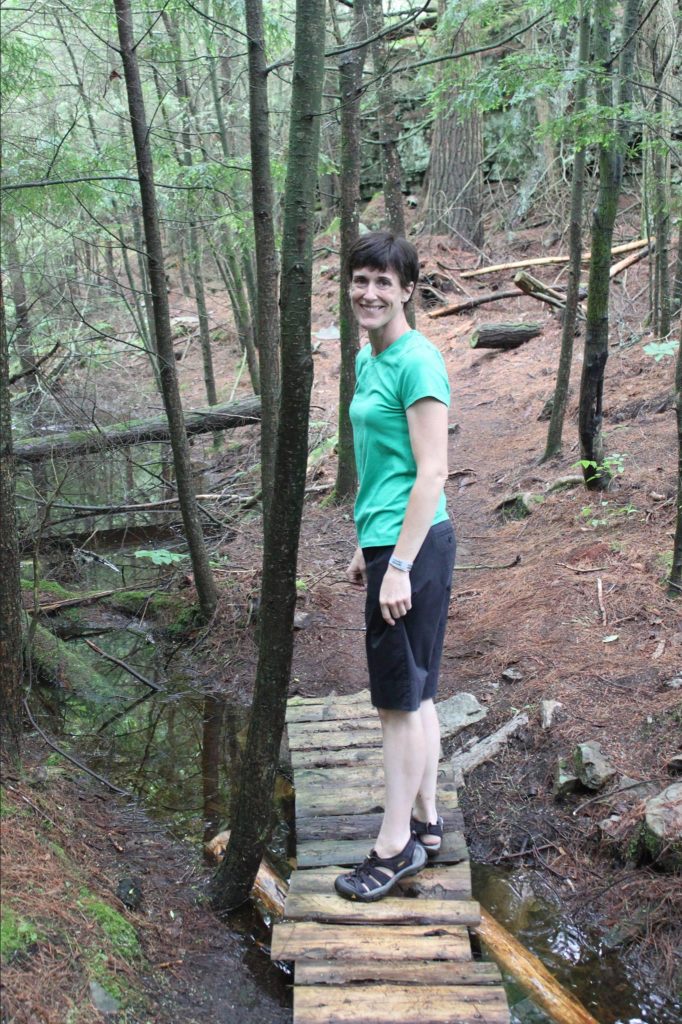 A photo of Claire hiking in the woods near her home in the Pocono Mountains. She's standing on a makeshift wooden bridge over a creek. Claire is wearing a short-sleeve shirt, black shorts, and lightweight hiking sandals. The trees are sending out green shoots, and the rocks by the creek are covered in moss.