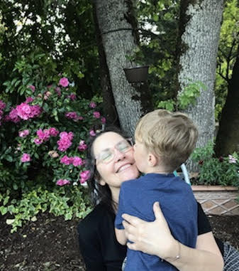 Lisa hugs her grandchild. There are trees and bushes in the background. Lisa crouches to get on the child's level; he looks to be 3 or 4 years old. Only the back of the child's blond head is visible.
