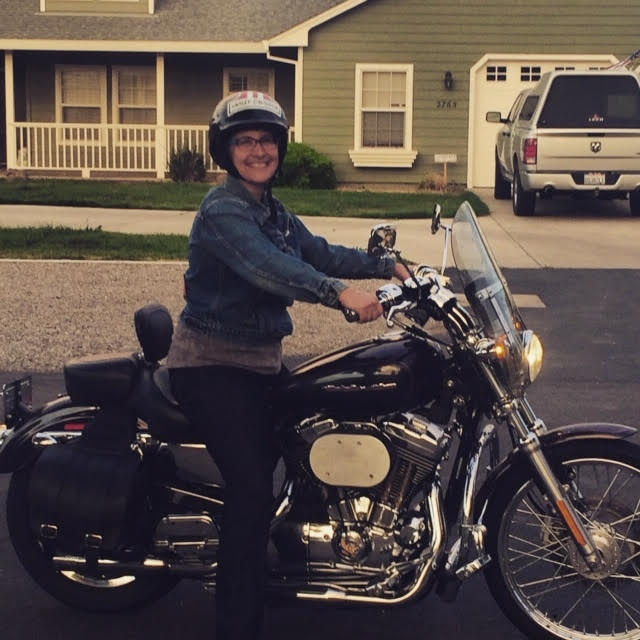 A photo of Lisa sitting on her Harley. A suburban house with a porch and garage are visible in the background. Lisa is wearing a helmet (maybe she finally learned), glasses, and a denim jacket. And a giant smile.