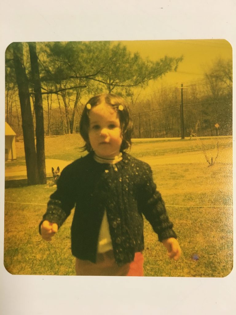 A faded photo of Claire, taken in 1972, when she was two years old. Like all toddlers, she seems to have little awareness of what's happening. She's outside, and a German shepherd is barely visible in the background behind Claire's left arm. She has two barrettes on either side of her forehead, and is wearing a dark sweater over a white shirt.