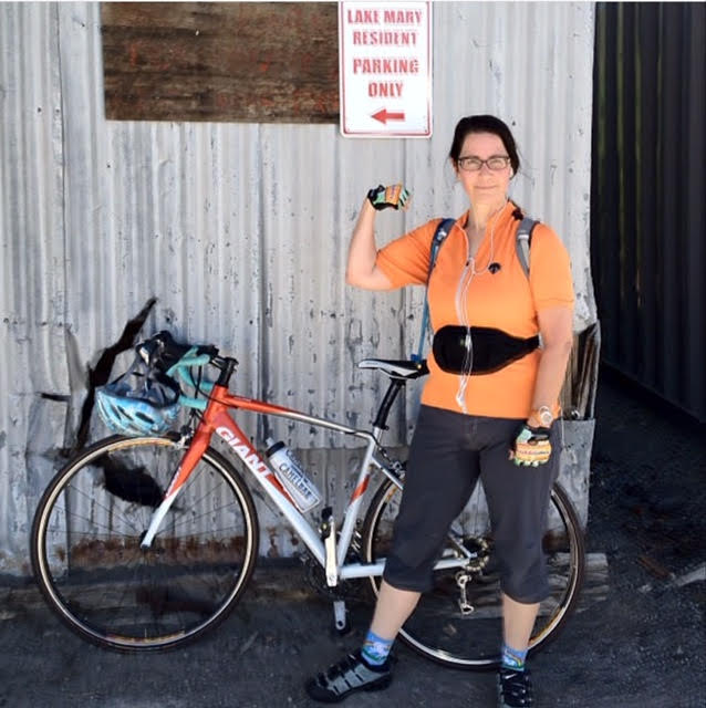 A photo of Lisa standing next to her bike in 2012. She's wearing a fanny pack, gloves, a backpack, and riding shorts, and is flexing to show off her biceps. A sign behind her reads, "LAKE MARY RESIDENT PARKING ONLY" so I sure hope she is one.