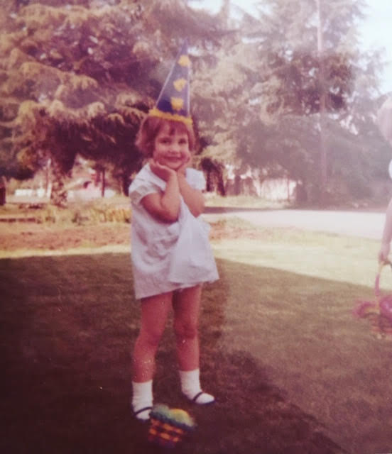 A photo of Lisa as a child. She's wearing a conical birthday hat, and has both hands under her chin. She's standing on a verdant lawn, and there are trees in the background.