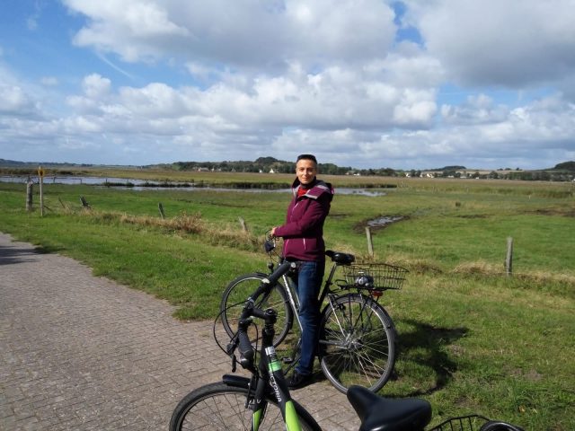 A photo of Francesca standing by her bike in front of a gorgeous landscape. Maybe Ireland? There are fluffy, cotton-ball clouds in the blue sky, and the pasture behind the road is green. Francesca's bike is parked on a tiled roadway. She's wearing jeans and a maroon jacket, and is looking at the camera.