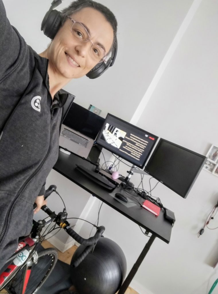 A selfie of Francesca, taken in front of her workstation. The photo was taken with the camera tilted about 45 degrees to the right. You can see the stationary bike Francesca uses when dialing into training meetings. An exercise ball is also visible under the desk.