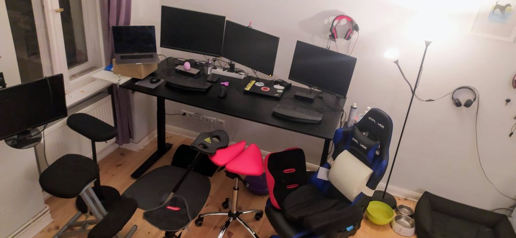 A photo of Francesca's messy workstation at home. On the left is an electronic arm with a monitor affixed. On the right is Francesca's desk, which supports a laptop, three computer monitors, and two keyboards. The desk itself is adjustable, and can be raised to a standing position. A collection of chairs are spread out in front of her desk. You can clearly make out the kneeling desk, saddle chair, and standard office chair with a collection of pads on top. Two pairs of headphones hang on the wall.