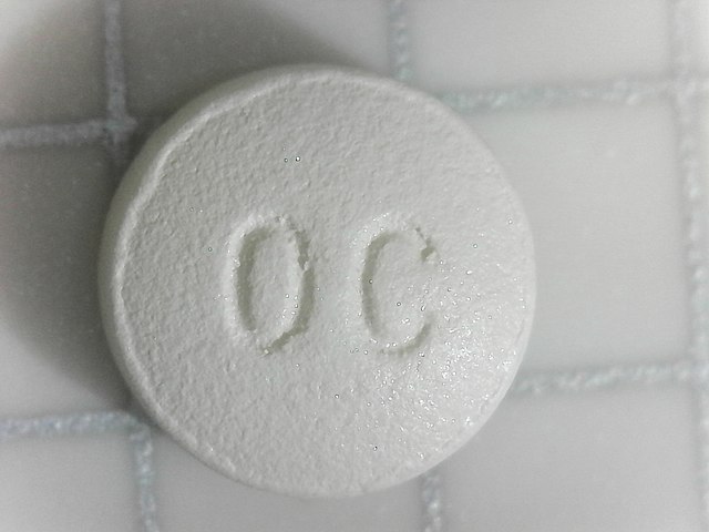 A photo of an OxyContin pill on a tile surface. The pill is round and white, and stamped with, "OC."