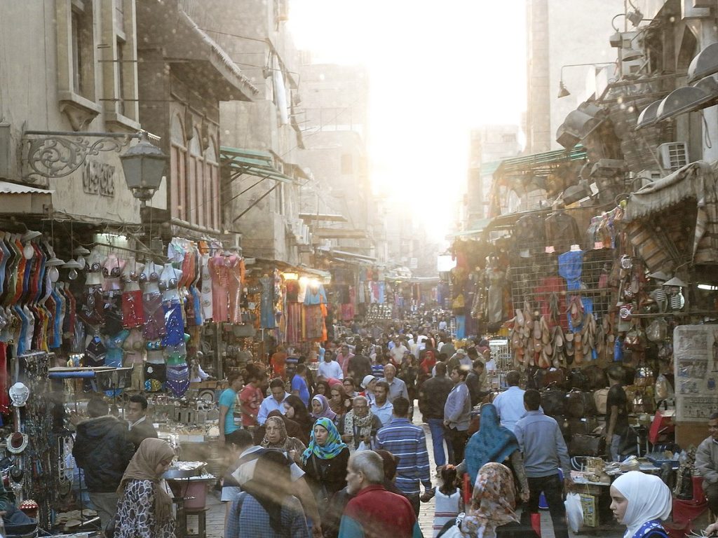 A photo of a crowded street in Cairo's Khan el-Khalili bazaar. Shoppers are milling around. Stalls selling clothing, jewelry, and souvenirs are set up on both sides of the street.