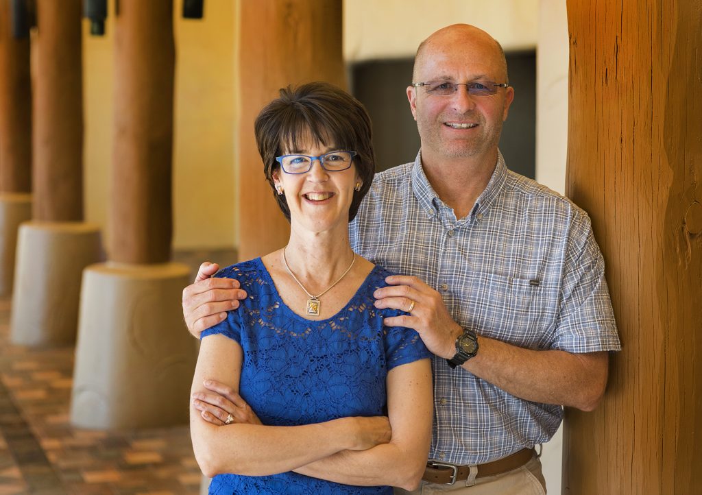 A photo of Linda and her husband Dale, taken in 2016. They are inside, though it's not clear where. There are wooden pillars visible in the background. The two of them are visible from the waist up. Linda is on the left. She has her arms crossed, and is smiling. Dale stands to her right, and his hands are placed on her shoulders. They both look happy.