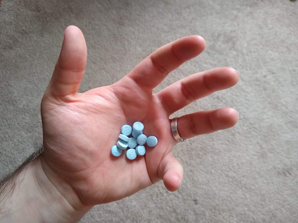 A close-up of a man's open hand. He is cupping a small pile of prescription antidepressant pills in his palm. My husband graciously agreed to serve as a hand model for this photo. Also, I stole all his pills, and dumped them all over the floor.