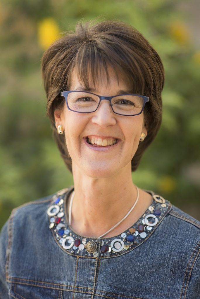 This headshot of Linda was taken in 2016. She has short, brown hair, blue wire-frame glasses, and a blue shirt with an embroidered collar. She's in her mid-fifties, and smiling.