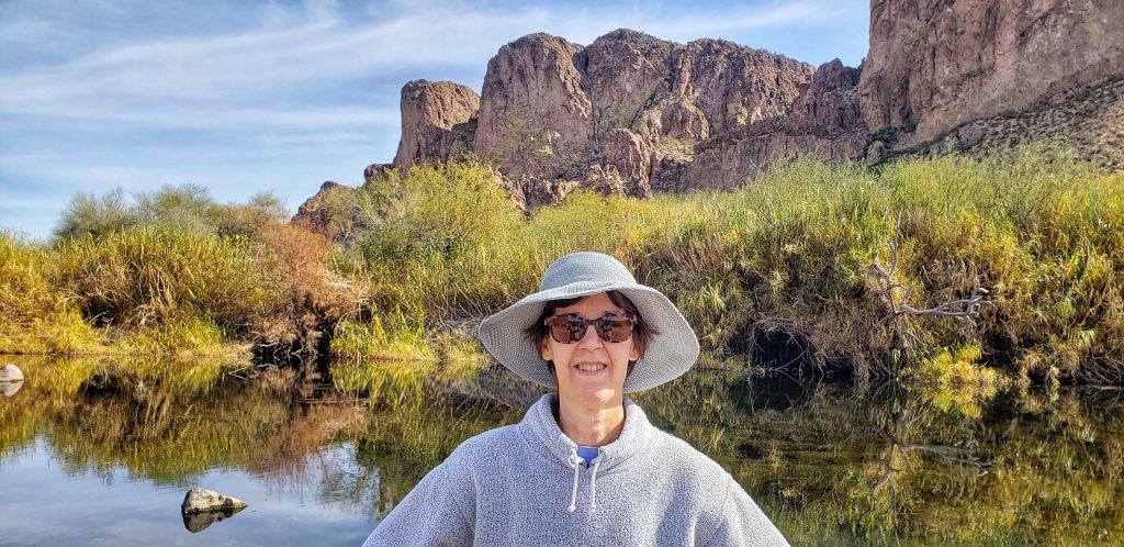 A photo of Linda, taken during her hike near Salt River. It must be a still day, because the water behind her is smooth as glass, and reflects the rocky shore and shrubs behind her. A butte rises up in the background.