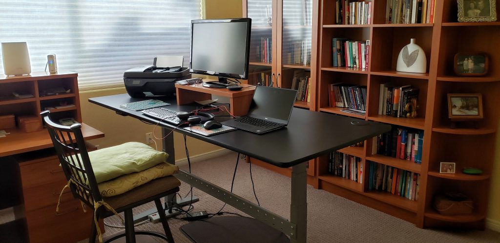 A photo of Linda's office, including her desk. The back wall of her office is covered in bookshelves, which hold books (duh). On top of the desk is a printer, computer monitor, keyboard, mouse, and laptop. The chair is a ladderback chair, rather than a standard office chair. There are several cushions stacked on top to make it softer. Linda also often uses ice packs to dull the pain.