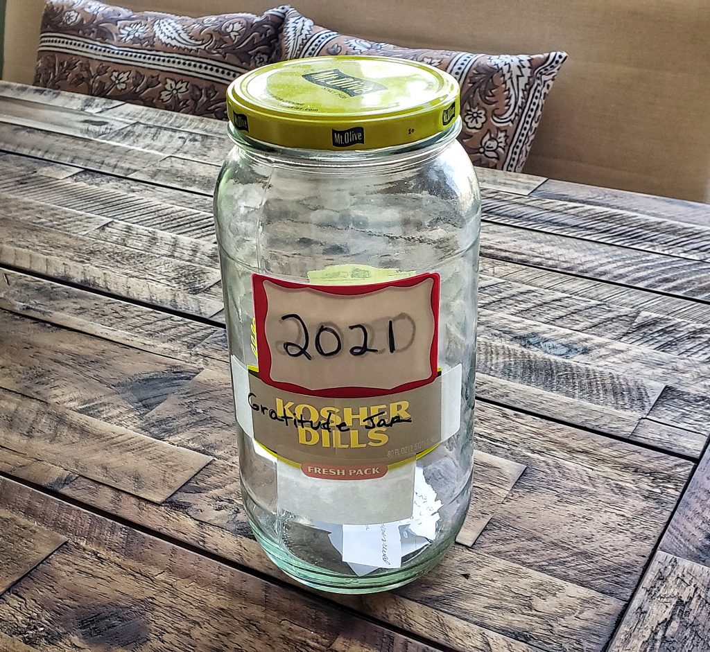 A photo of Linda's gratitude jar, placed on a wooden table. The jar is an old pickle jar. Each year, she puts a new label on the jar with the current year written on it. You can see the layers of labels. Since this photo was taken in January, there are only a few slips of paper in the jar.