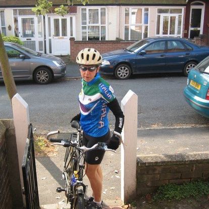 This photo shows Julie standing beside her bike during in Norfolk in 2008. She is passing through a low gateway into the yard. The street behind her is lined with cars, and other houses are visible on the other side. Julie is wearing a helmet, sunglasses, athletic jacket, and gloves. She looks like she either just finished kicking ass, or is about to.