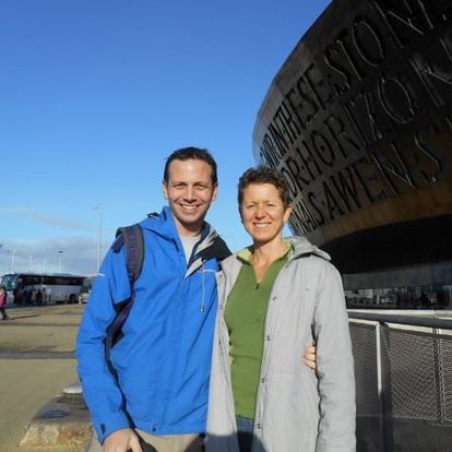 Paul and Julie stand in front of the Wales Millenium Center in Cardiff, Wales. You can tell they're not in the tropics anymore. It's a sunny day, and the two are dressed in jackets that look waterproof.