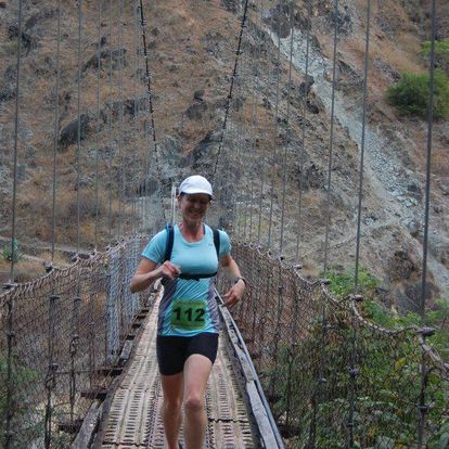This photo of Julie, taken in 2012, shows her in her first Skyrun in Manilla. A mountain covered in reddish scrub is rising up behind her. Julie appears to running across some sort of a bridge, with wires on both sides. She's wearing shorts, a baseball cap, and "112" race number.