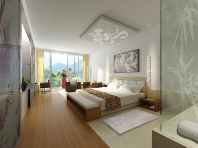 This photo shows the interior of a luxurious hotel room which I would love to stay at, standing desk or no. The floor is made of bleached wood, the walls are white, and the carpets and trim are cream and white. There are abstract paintings on the ceiling and wall above the bed. On the far wall, there's a sliding glass door and large windows. Two women are standing out on the balcony, staring at the forested mountains in the distance. There are two chairs and a couch inside the door, and a huge bed neatly made.