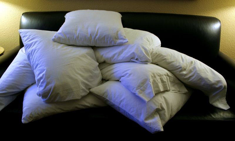 This photo shows a pile of white bed pillows that threaten to subsume the black couch beneath it. The scene is illuminated with two lamps off to each side.