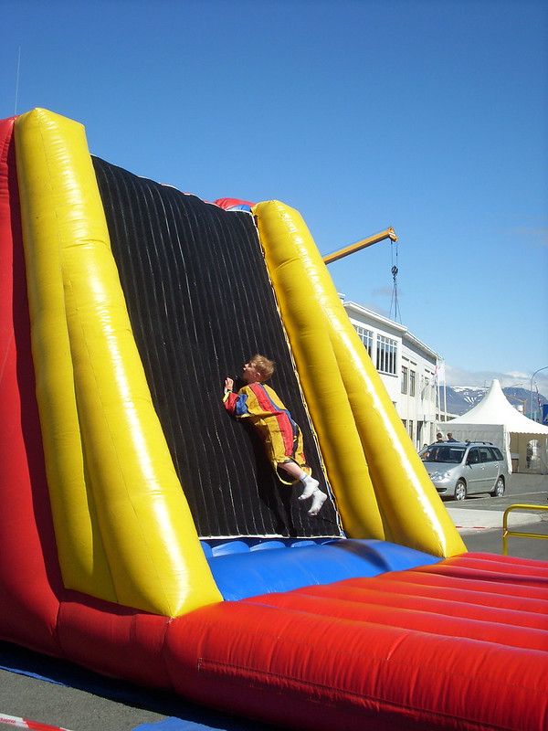 This photo was taking during a sunny day at a harbor. There is a large, inflatable bounce pad and wall. The wall is covered with Velcro. A boy of about 10 is seen wearing a Velcro suit. He's rebounding off the jump pad, and heading straight for the wall.