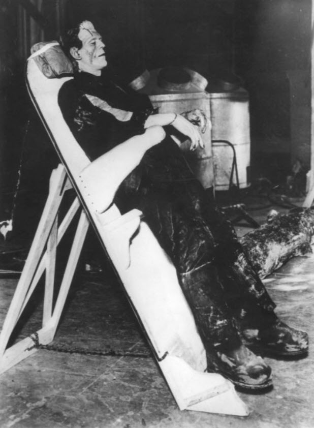 A black and white image of Boris Karloff in full Frankenstein makeup, reclining on his wooden gurney. The gurney is shaped sort of like the lowercase Greek letter lambda. The part Boris leans on is tilted about 20 degrees back, and a kickstand sort of thing holds it up from behind.