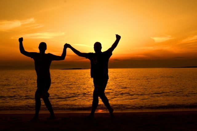 This photo shows the silhouettes of two people walking on the beach at sunset. They're holding hands, and raising their arms in a jubilant pose. The features of the people aren't distinctly visible. I think they're both men? The sky and water behind them are a lovely golden orange color.