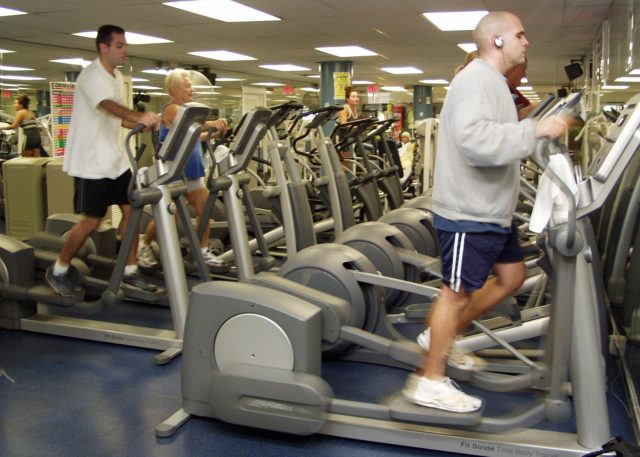 A photo of the inside of a gym. Fluorescent lights shine down on the two rows of elliptical machines. Several people are striding away on them. The man in the foreground, on the right, is striding along determinedly while wearing small headphones. He's wearing a sweatshirt and shorts.
