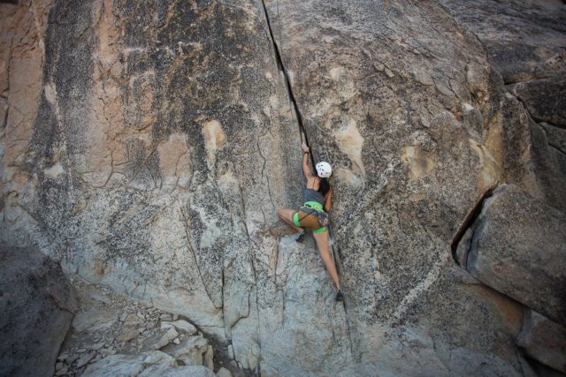 A female rock climber is scaling the face of a cliff. She's wearing a helmet, shorts, and a bright green harness with three loops that wrap around her waist and thighs. The rope she's tied to is barely visible.